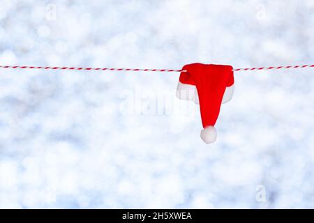 Eine kleine rote Weihnachtsmütze hängt an einem Seil auf einem verschwommenen blau-grauen Hintergrund. Speicherplatz kopieren. Leer für Design, Konzept Stockfoto