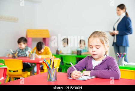 Nettes präteen Mädchen studieren im Klassenzimmer auf Hintergrund mit Klassenkameraden und Lehrer Stockfoto
