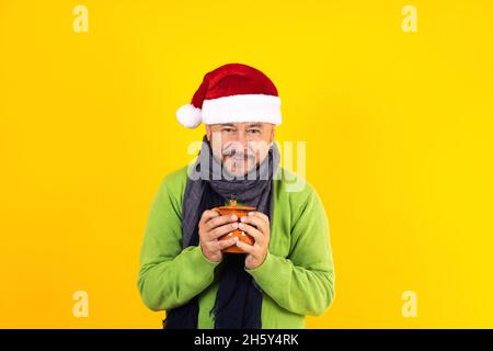 hispanischer älterer Mann mit einem Tonkrug Kaffee oder Fruchtpunsch zu weihnachten auf gelbem Hintergrund in lateinamerika Stockfoto