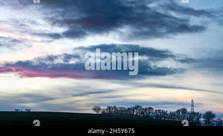 In schönem, sanft rollendem Ackerland, übersäten blattlosen Bäumen auf winterlichem Ackerland in Südengland, geschichteten Wolken, die rot mit Nachlicht leuchten Stockfoto