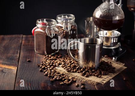 Metallische Tasse und Vakuum-Kaffeemaschine auch bekannt als vac-Topf, Siphon oder Syphon-Kaffeemaschine und geröstete Kaffeebohnen auf rustikalem Holztisch Stockfoto