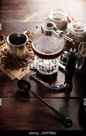 Vakuum-Kaffeemaschine auch als vac-Topf, Siphon oder Syphon Kaffeemaschine bekannt. Metallbecher und geröstete Kaffeebohnen auf rustikalem Holztisch Stockfoto