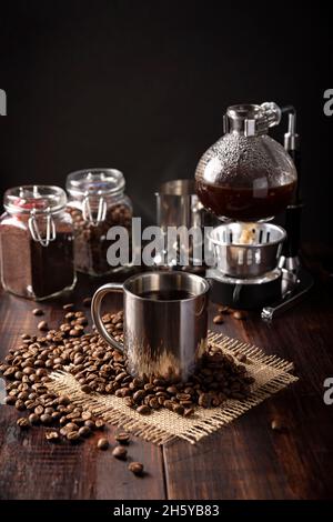 Metallische Tasse und Vakuum-Kaffeemaschine auch bekannt als vac-Topf, Siphon oder Syphon-Kaffeemaschine und geröstete Kaffeebohnen auf rustikalem Holztisch Stockfoto