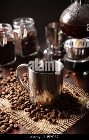 Metallische Tasse und Vakuum-Kaffeemaschine auch bekannt als vac-Topf, Siphon oder Syphon-Kaffeemaschine und geröstete Kaffeebohnen auf rustikalem Holztisch Stockfoto