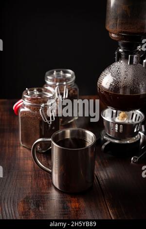 Metallische Tasse und Vakuum-Kaffeemaschine auch bekannt als vac-Topf, Siphon oder Syphon Kaffeemaschine und Kaffeeglasbehälter auf rustikalem Holztisch Stockfoto