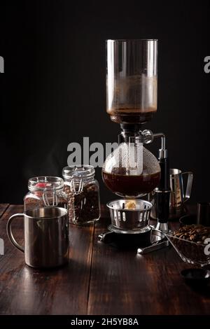 Vakuum-Kaffeemaschine auch als vac-Topf, Siphon oder Syphon Kaffeemaschine bekannt. Metallbecher und geröstete Kaffeebohnen auf rustikalem Holztisch Stockfoto