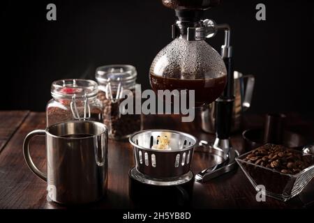 Vakuum-Kaffeemaschine auch als vac-Topf, Siphon oder Syphon Kaffeemaschine bekannt. Metallbecher und geröstete Kaffeebohnen auf rustikalem Holztisch Stockfoto