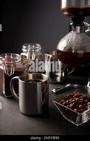 Vakuum-Kaffeemaschine auch als vac-Topf, Siphon oder Syphon Kaffeemaschine bekannt. Metallbecher und geröstete Kaffeebohnen auf rustikalem Tisch aus schwarzem Stein. Stockfoto