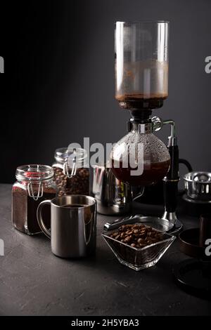 Vakuum-Kaffeemaschine auch als vac-Topf, Siphon oder Syphon Kaffeemaschine bekannt. Metallbecher und geröstete Kaffeebohnen auf rustikalem Tisch aus schwarzem Stein Stockfoto