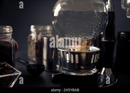 Nahaufnahme der Vakuumkaffeemaschine mit Feuerzeugheizung, auch bekannt als vac-Kaffeemaschine, Siphon oder Syphon auf rustikalem Tisch aus schwarzem Stein. Stockfoto