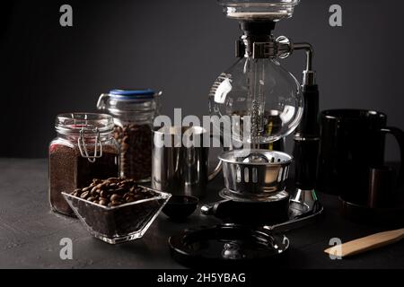 Vakuum-Kaffeemaschine auch als vac-Topf, Siphon oder Syphon Kaffeemaschine bekannt. Metallbecher und geröstete Kaffeebohnen auf rustikalem Tisch aus schwarzem Stein. Stockfoto