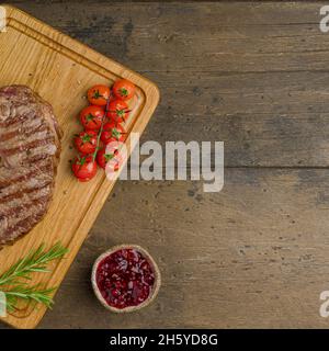 Gebratenes Striploin-Steak mit Kirschtomaten, Thymian, Rosmarin und Preiselbeersoße auf Holzschneidebrett. Köstliches saftiges und zartes Fleisch. Overhead, Stockfoto
