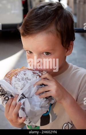 Das Kind isst eine große runde Brötchen. Porträt des Kindes, das ein Brioche-Brötchen mit Puderzucker beißt Stockfoto