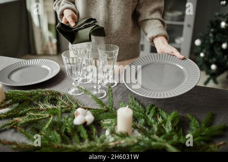 Nahaufnahme einer jungen Frau, die Teller auf dem Esstisch aufstellt, der zu Weihnachten mit Tannenzweigen und Kerzen in Grautönen dekoriert ist, Kopierraum Stockfoto