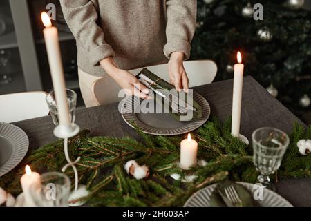 Nahaufnahme einer nicht erkennbaren Frau, die zu Weihnachten einen Esstisch mit Tannenzweigen und Kerzen in Grautönen eingerichtet hat, Kopierraum Stockfoto