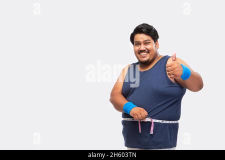 Porträt eines fetten Mannes, der lächelt und die Daumen hoch zeigt, während er vor schlichtem Hintergrund ein Maßband um seine Taille wickelt. Stockfoto