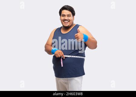 Porträt eines fetten Mannes, der lächelt und die Daumen hoch zeigt, während er vor schlichtem Hintergrund ein Maßband um seine Taille wickelt. Stockfoto