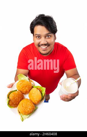 Porträt eines fetten Mannes, der vor einfachem Hintergrund ein Getränk und einen Teller Burger in der Hand hält. Stockfoto