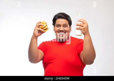 Porträt eines fetten Mannes, der vor einfachem Hintergrund ein Getränk und einen Burger in der Hand hält. Stockfoto