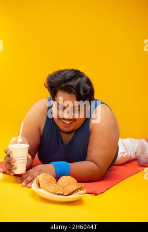 Ein fetter Mann, der auf einer Yogamatte liegt, hält ein Getränk gierig auf dem Teller des Burgers. Stockfoto