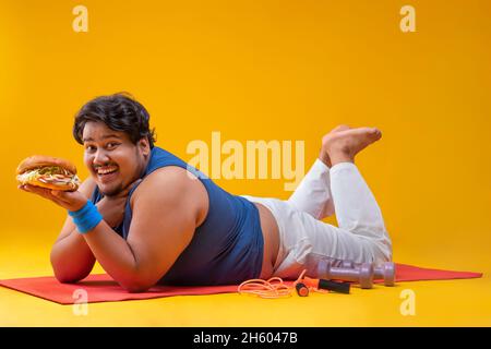 Ein fetter Mann, der auf einer Yogamatte liegt und einen Burger mit Dummeln hält und ein Springseil neben sich hält. Stockfoto