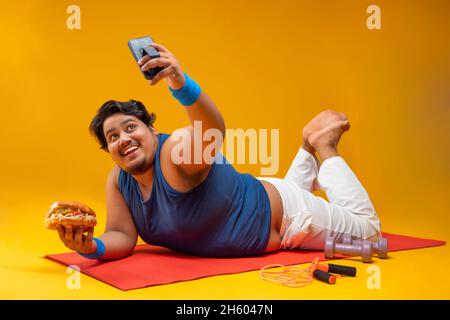 Ein fetter Mann, der auf einer Yogamatte liegt, einen Burger hält und mit Dummeln auf ein Selfie klickt und dabei ein Springseil hält. Stockfoto