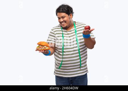 Porträt eines fetten Mannes mit Maßband um den Hals, der gierig einen Burger verschlingt, anstatt einen Apfel. Stockfoto