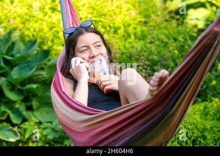 Wunderschöne Teenager-Mädchen auf der Hängematte liegend entspannen, trinken Orangensaft und mit dem Smartphone Stockfoto