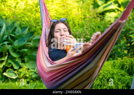 Wunderschöne Teenager-Mädchen auf der Hängematte liegend entspannen, trinken Orangensaft und mit dem Smartphone Stockfoto