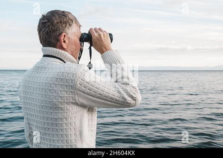 Porträt eines hübschen Mannes mittleren Alters in einem weißen Pullover, der durch ein Fernglas auf das Meer blickt. Speicherplatz kopieren. Stockfoto