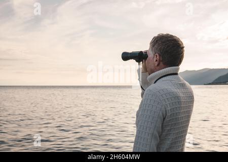 Porträt eines hübschen Mannes mittleren Alters in einem weißen Pullover, der am Abend bei Sonnenuntergang durch ein Fernglas auf das Meer blickt. Platz für Text. Stockfoto