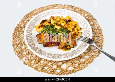 Stücke von gebackenem Schweinefleisch, geschmortem Spinat und gerösteten Gnocchi (Knödel) auf Teller auf kreisförmigen Bambuskissen auf weißem Hintergrund, isoliert, Nahaufnahme. Stockfoto