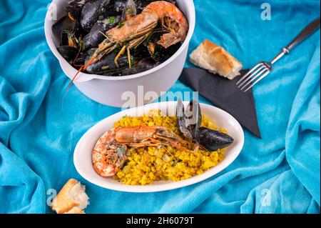 Meeresfrüchte auf blauem Tisch. Kleiner ovaler Teller mit traditioneller Paella (Vorspeise) und großer Schüssel mit schwarzer Muschel und Garnelen, Gabel und Brot. Stockfoto