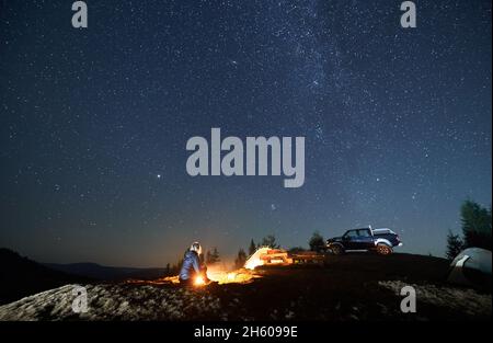 Einsame Frau sitzt auf Gras am brennenden Feuer in der Nähe von zwei Zelten und schwarzem SUV. Nächtliches Campen auf einem Berg unter Sternenhimmel. Baumkronen und Silhouetten von Bergen weit im Hintergrund. Stockfoto