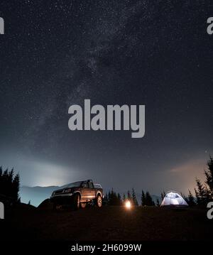 Farbenprächtiger Sternenhimmel am Abend und Milchstraße über dem Ferienlager auf einem Berghang. Schwarzes SUV, helles Zelt und Lagerfeuer zwischen ihnen auf dem Hintergrund von Silhouetten von Bäumen und Bergen. Stockfoto