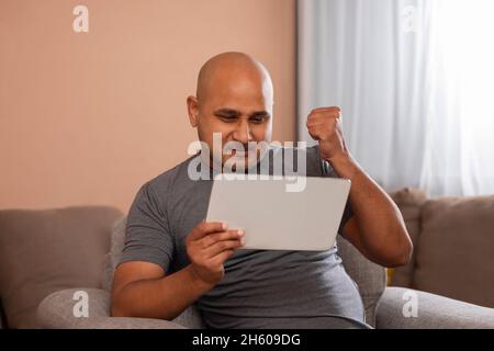 Ein Glatzkopf, der mit der Faust den Sieg zelebriert, während er auf einem digitalen Tablet in der Hand zuschaut. Stockfoto