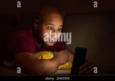 Ein kahler Mann, der im Dunkeln etwas auf dem Handy beobachtet, mit erstaunlicher Gesichtsausdruck. Stockfoto