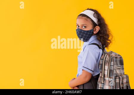 EIN MÄDCHEN IN SCHULUNIFORM, DAS ZUR SCHULE GEHT, WÄHREND ES DIE KAMERA MIT EINER GESICHTSMASKE ANSCHAUT Stockfoto