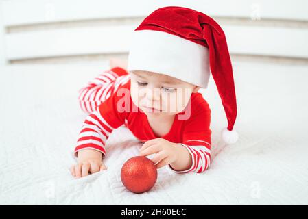 Ein einjähriges Baby im Weihnachtsmütze liegt auf dem Bauch und spielt mit scharlachroten Weihnachtsball. Kinder tragen einen rot-weiß gestreiften Anzug. Frohe Weihnachten und glücklich Stockfoto