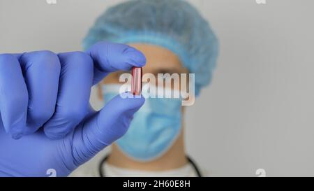 Ein hübscher junger Arzt untersucht eine neue Droge. Ein junger, gutaussehender Arzt in einer medizinischen Maske hält eine rote Pille in seinen Händen und präsentiert eine neue Droge. Stockfoto