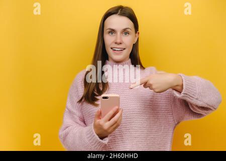 Überrascht überrascht junge Brünette Frau trägt rosa gestrickt Pullover stehen Zeigefinger auf Handy Eingabe sms-Nachricht Stockfoto