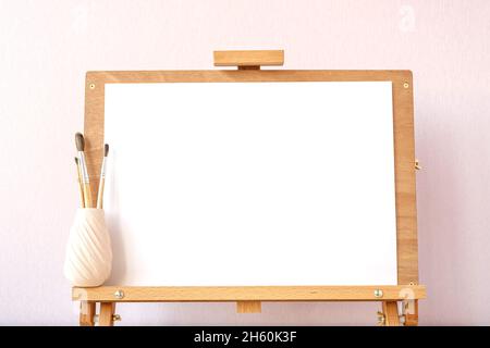 Leeres weißes Papiermockup zum Malen auf hölzerner Staffelei auf Tisch im Heimstudio Stockfoto