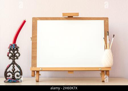 Leeres weißes Papier zum Malen auf HolzStaffelei, Pinsel und Kerze auf dem Tisch im heimischen Atelier Stockfoto