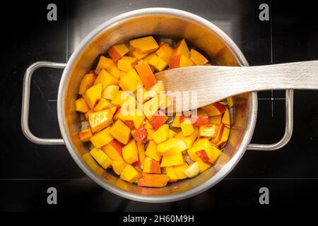 Roter Kürbis oder Hokkaido-Kürbis in Scheiben in einem Edelstahltopf auf einem schwarzen Herd, beim Kochen einer herbstlichen Gemüsemahlzeit, Blick von oben, Stockfoto