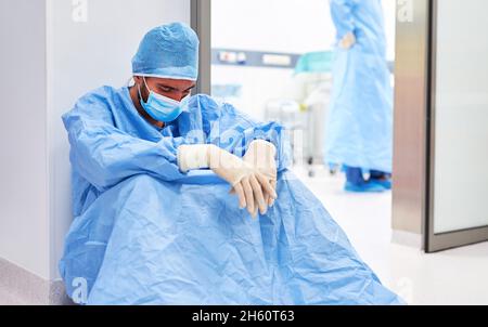 Erschöpfter Chirurg oder Krankenschwester in Schutzkleidung sitzt ausgebrannt vor dem Operationssaal Stockfoto