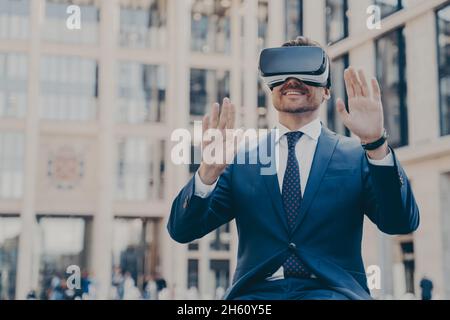 Junger, glücklicher bärtiger Büroangestellter, der formell im Freien sitzt und VR-Headsets oder 3d-Brillen verwendet Stockfoto
