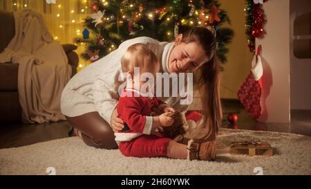 Junge lächelnde Mutter, die ihrem kleinen Sohn im Weihnachtsmann-Kostüm neben dem Weihnachtsbaum einen warmen Stiefel zukleidet. Familien und Kinder feiern den Winter Stockfoto