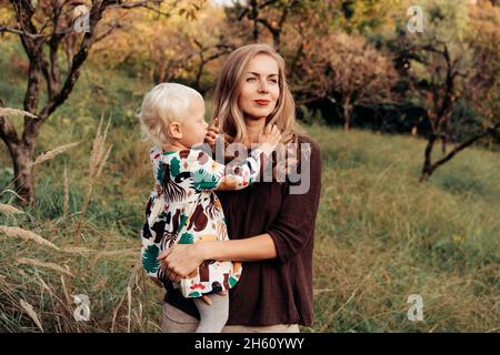 Eine charmante Mutter hält die Tochter des Kleinkindes während eines Herbstspaziergangs in der Natur in den Armen. Stockfoto