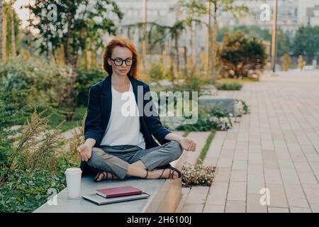 Junge entspannte Rotschopf Frau meditiert im Park fühlt sich ruhig sitzt in Lotus-Pose im Freien Stockfoto
