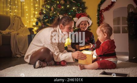 Glückliche Mutter mit Baby und älterem Sohn, der Geschenkboxen neben dem Weihnachtsbaum im Haus präsentiert. Familien und Kinder feiern Winterferien. Stockfoto